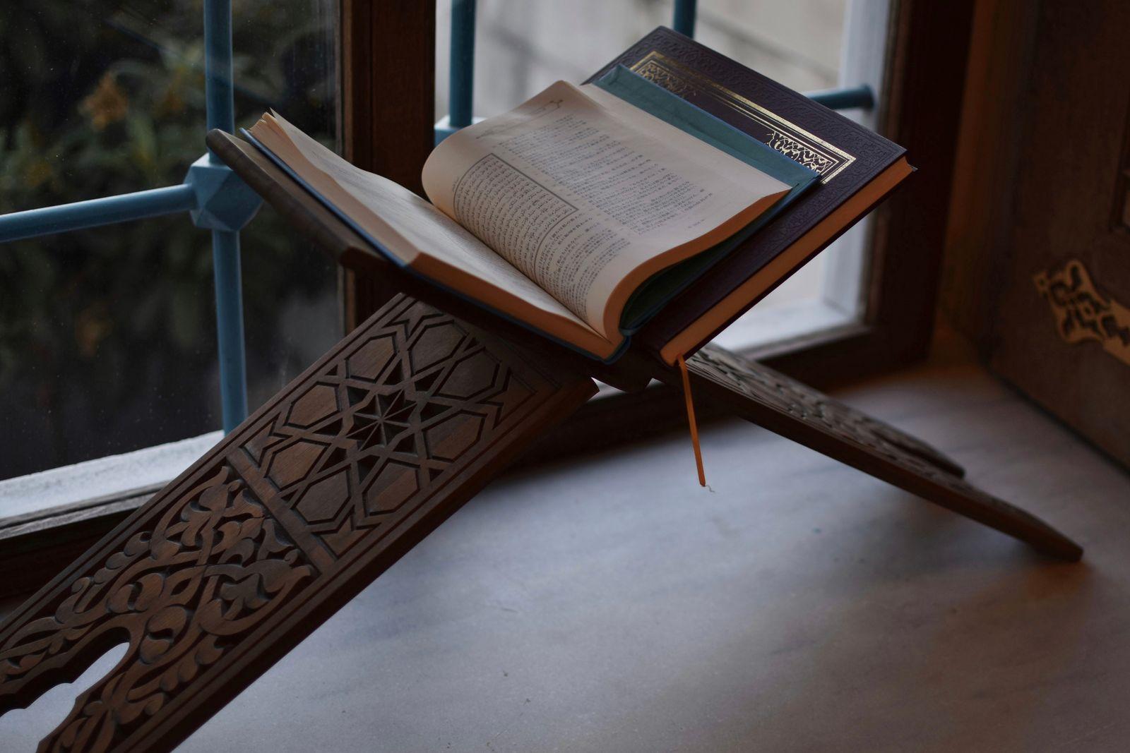 An open Quran resting on a carved wooden rehal stand by a window